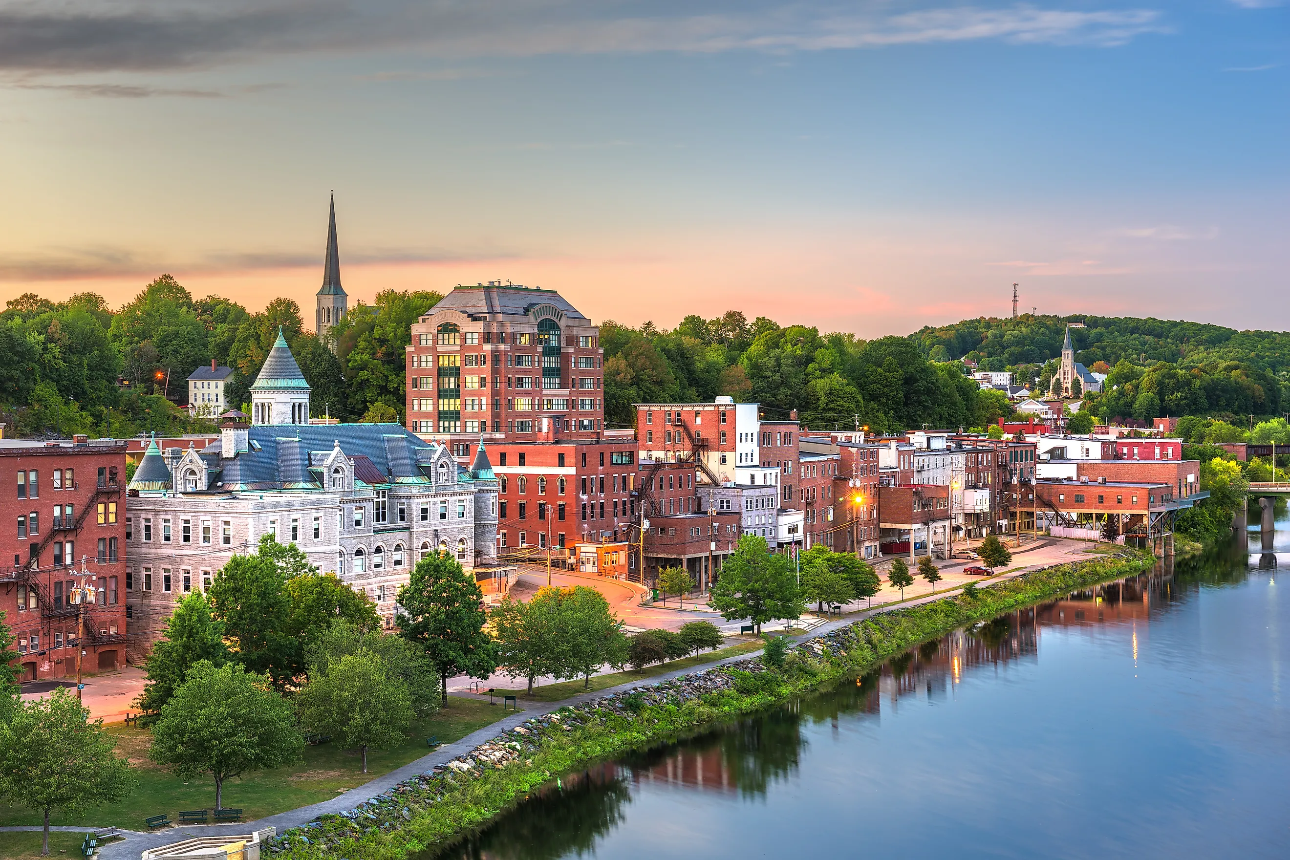A beautiful sunset view of Augusta, Maine, showcasing the historic downtown district, Kennebec River waterfront, classic New England architecture, church steeples, brick buildings, and scenic riverside landscape.